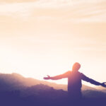 Osoba stojaca na kopci s rozpaženými rukami, obrátená k vychádzajúcemu slnku, symbolizuje nový začiatok a slobodu.