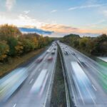 Diaľnica s hustou premávkou, na ktorej sa pohybujú autá a nákladné vozidlá vysokou rýchlosťou. Na oboch stranách cesty sú stromy s jesenným lístím a obloha sfarebnená západom slnka.