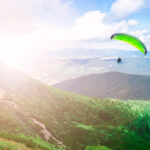 Paraglajdista na krídle padáka letiaci vysoko nad kopcami počas slnečného jarného dňa.